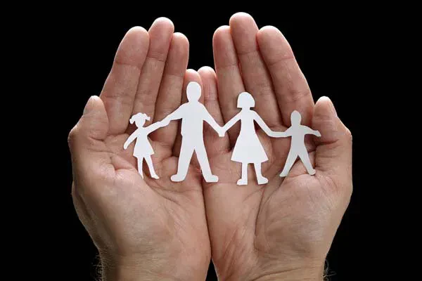 Two hands gently cup paper cutouts of a family with two adults and two children, symbolizing protection, care, and unity, against a black background.