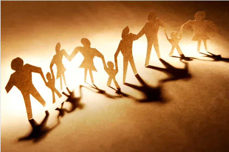 Paper cutouts of people holding hands in a line, casting shadows on a warm-toned surface, symbolizing unity, family, or community.
