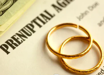 Two gold wedding bands resting on a document titled "PRENUPTIAL AGREEMENT," with a stack of US dollar bills partially visible in the top left corner.