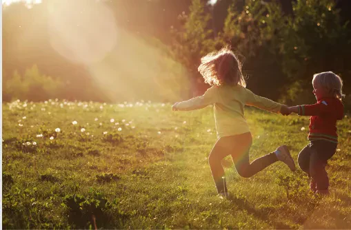 An image of two young children, possibly a girl and a boy, holding hands and running joyfully through a sun-drenched grassy meadow during a golden hour sunset, with warm sunlight creating a beautiful lens flare effect in the background.