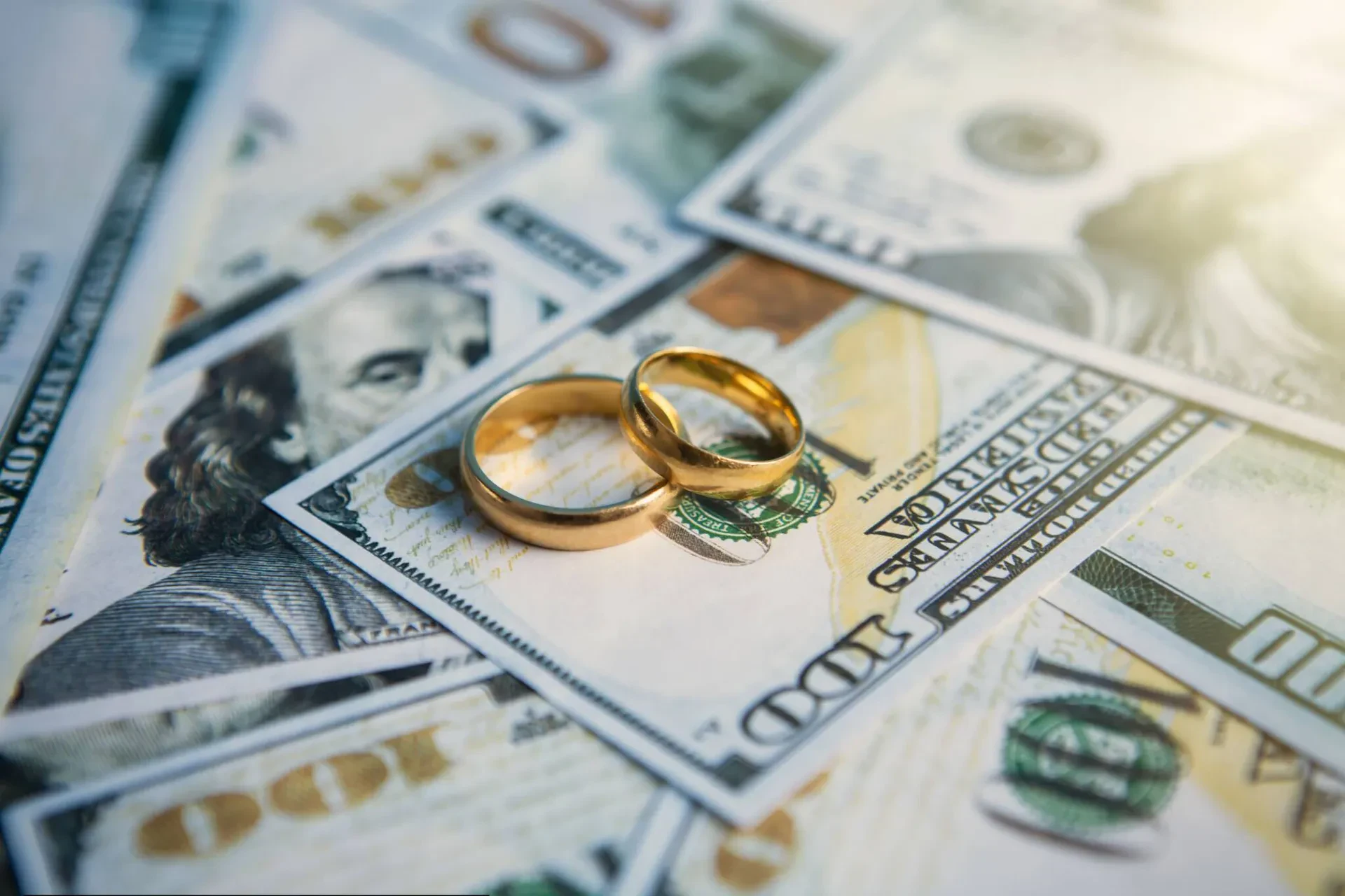 Two gold wedding bands resting on a document titled "PRENUPTIAL AGREEMENT," with a stack of US dollar bills partially visible in the top left corner.