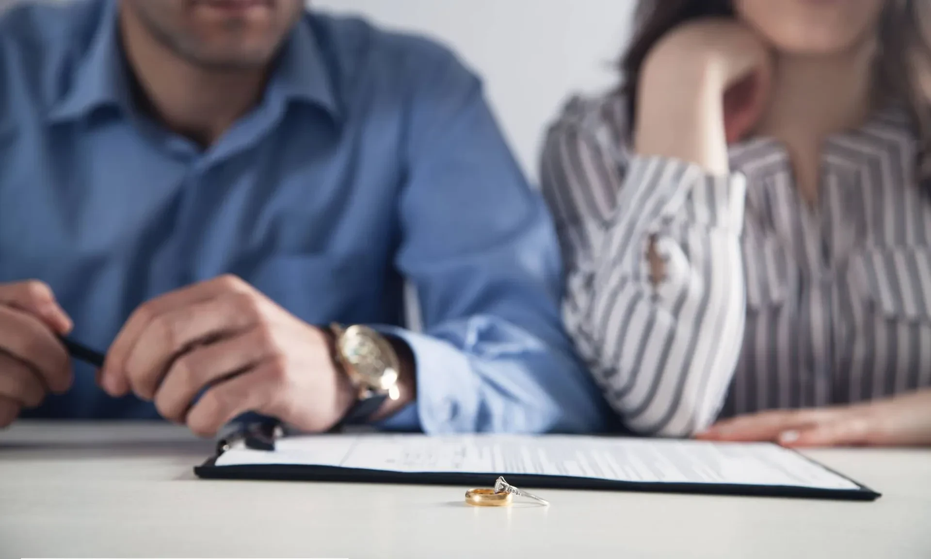 A man and woman sit at a table with a document and pen in front of them. Two rings are placed on top of the document, suggesting a legal or relationship decision. Their faces are only partially visible.