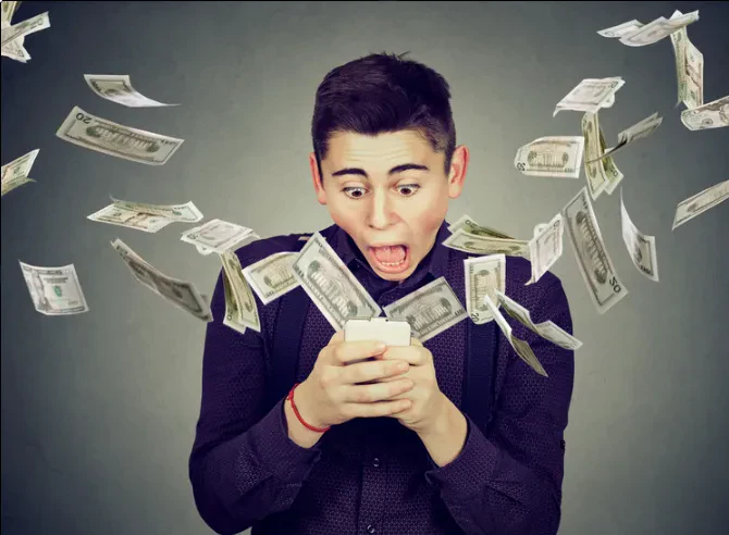 A young man with wide eyes and an open mouth stares in shock at his smartphone as dollar bills fly out from the screen and swirl all around him.
