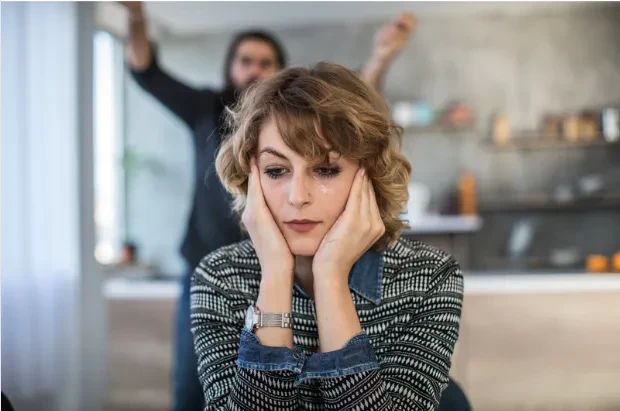A woman sits with her head in her hands, looking sad with a tear on her cheek, while someone gestures emphatically in the blurred background.