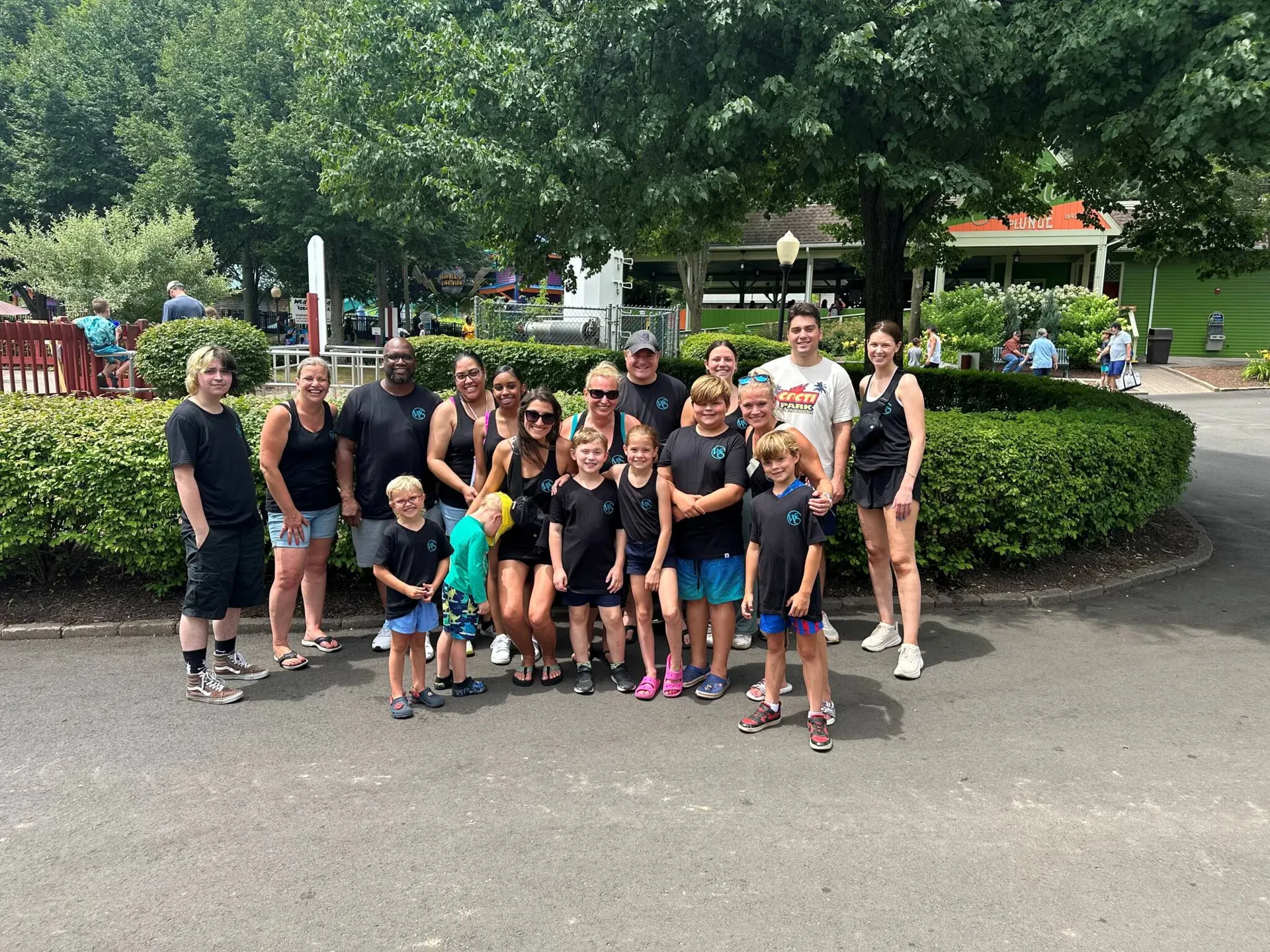 A group of 18 people, including adults and children, pose together outdoors on a paved path with greenery and trees in the background, all smiling at the camera.
