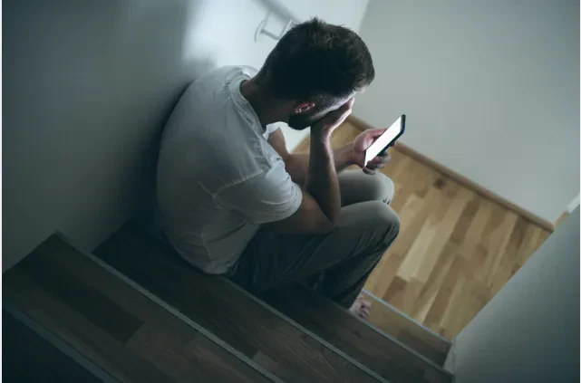 A man sits on stairs, holding his phone and resting his head in his hand, appearing stressed or upset