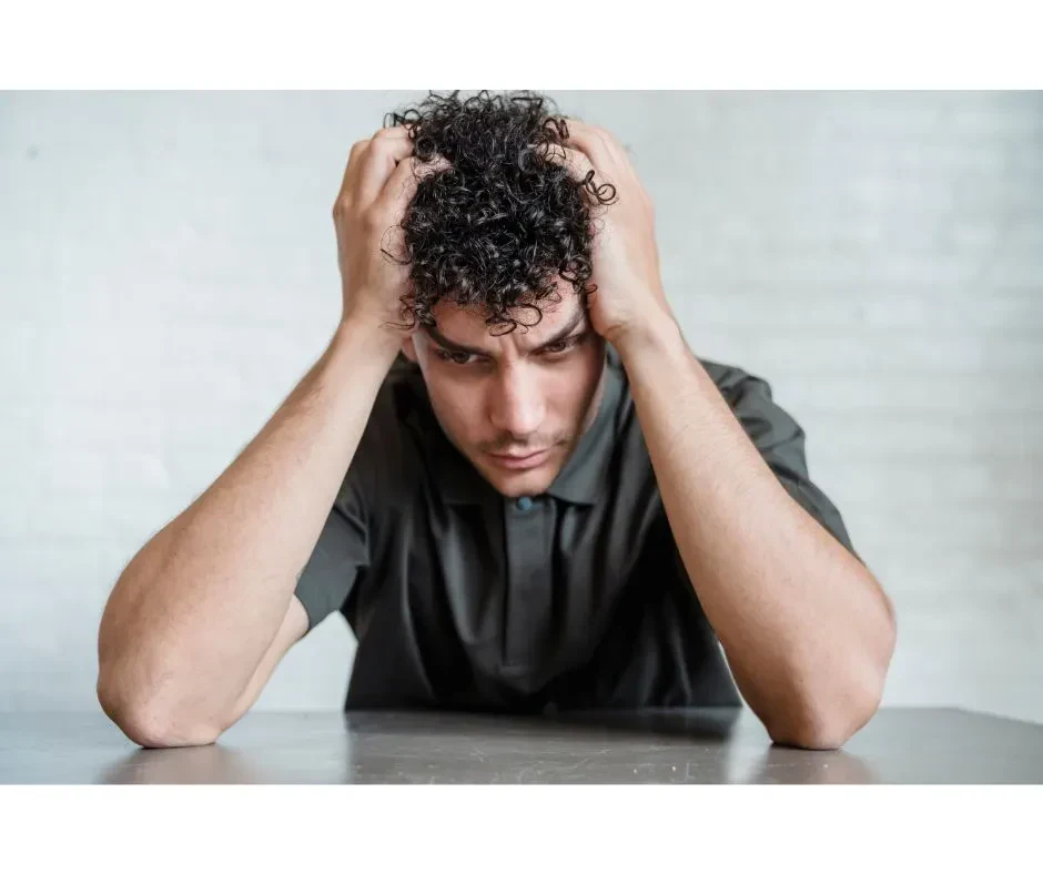 A man with curly dark hair sits at a table, holding his head in his hands with a stressed and worried expression.