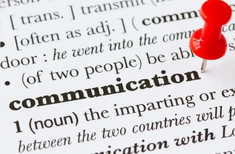 Close-up of a printed dictionary page focusing on the bolded word "communication," with a bright red pushpin stuck into the paper directly to its right.