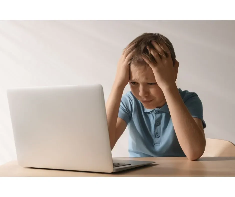 A boy sits at a desk with his head in his hands, staring at a laptop, conveying a sense of frustration or stress.