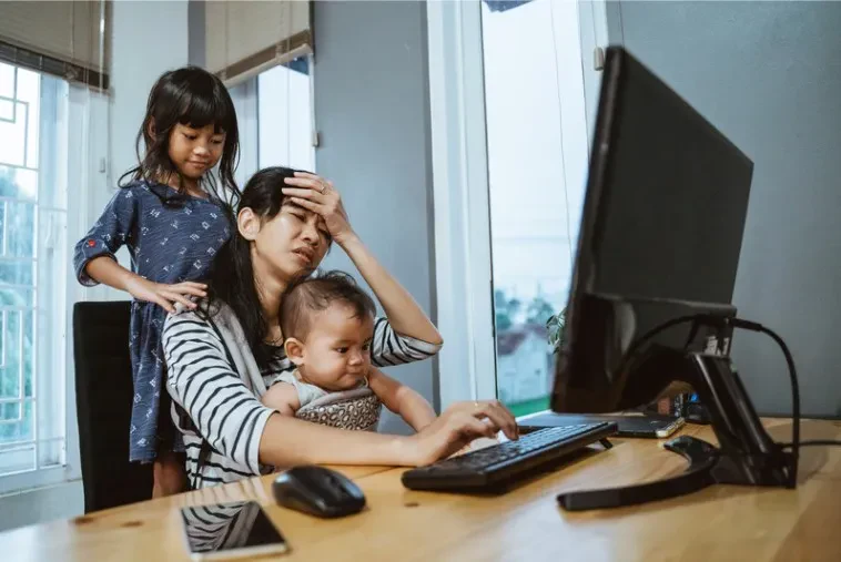 A woman sits at a wooden desk with a computer monitor, keyboard, and mouse, looking deeply stressed with her hand on her forehead while trying to type. She holds a young baby in her lap, while an older girl stands behind her chair, gently touching her shoulder.