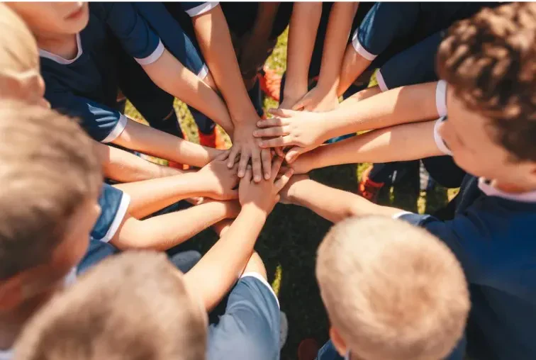 A group of children in sports uniforms stand in a circle, stacking their hands together in the center, showing teamwork and unity.