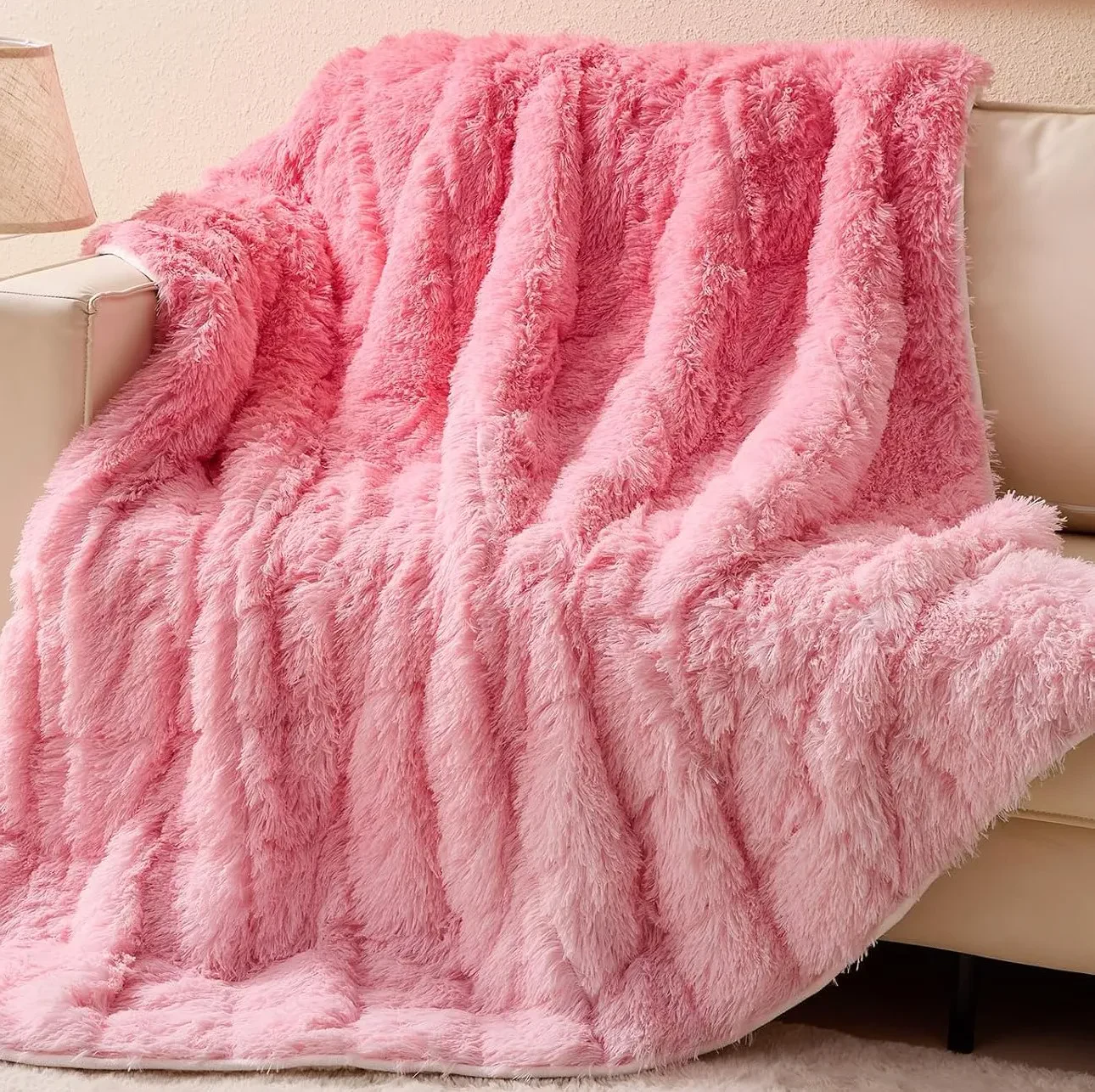 A fluffy, pastel pink faux fur throw blanket draped over the arm and cushions of a cream-colored sofa.