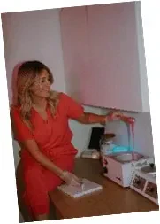 A woman in an orange outfit sits in front of a microwave, looking thoughtfully at the appliance.