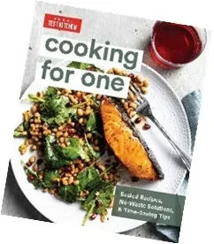 An image of the book cover for "Cooking for One" by America's Test Kitchen, featuring a plated meal with a seared salmon filet, a black-eyed pea and spinach salad, and a glass of red wine.
