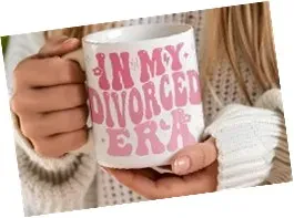 An image of a white ceramic mug being held by two hands. The mug features retro, wavy pink text that reads "IN MY DIVORCED ERA" surrounded by small pink sparkles and stars.