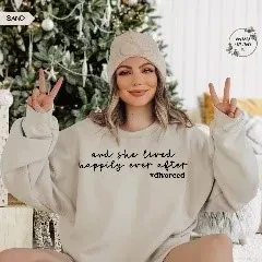 A woman smiling and flashing peace signs with both hands, wearing a light beige knit beanie and a matching sand-colored oversized sweatshirt that reads 'and she lived happily ever after divorced'. She is sitting indoors in front of a decorated Christmas tree.