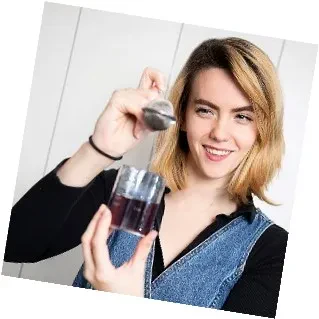 A woman holds a glass of liquid in one hand and a spoon in the other, smiling at the camera.