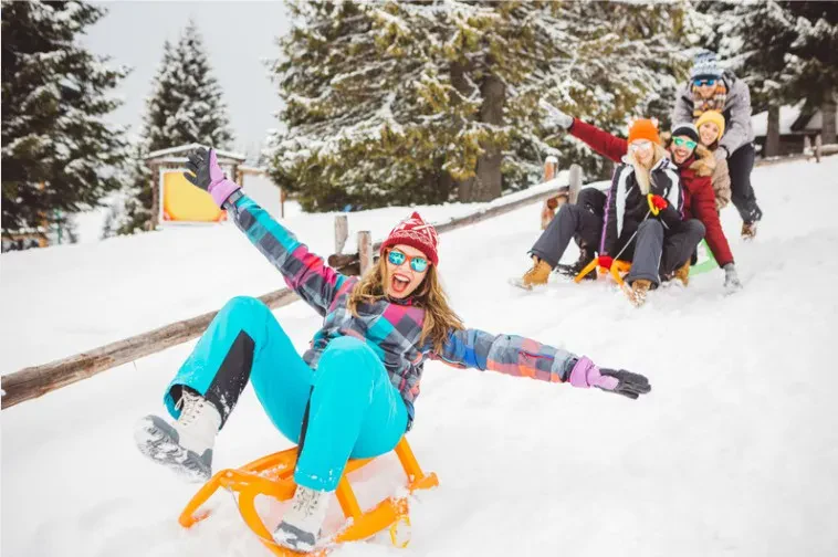 A group of people joyfully sledding down a snowy hill, surrounded by trees and a clear blue sky.