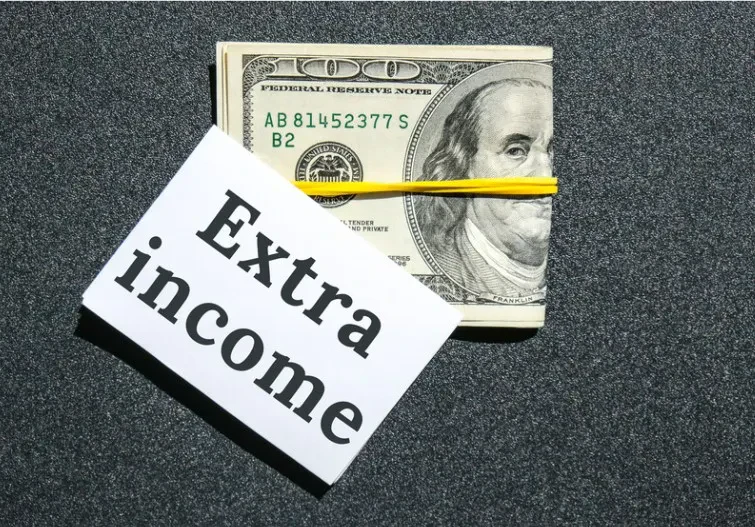 A stack of $100 bills secured with a yellow rubber band sits on a dark surface next to a white paper labeled Extra income.