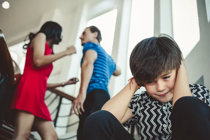 A boy sits on the stairs, covering his ears as his parents argue in the background.