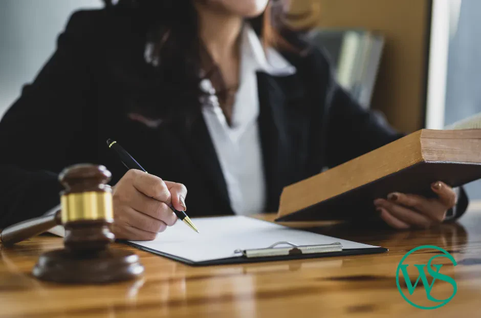 A person in a suit writes on a clipboard with one hand and holds a book in the other. A gavel sits on the wooden desk, and a green WS logo is in the lower right corner. The persons face is partially out of frame.