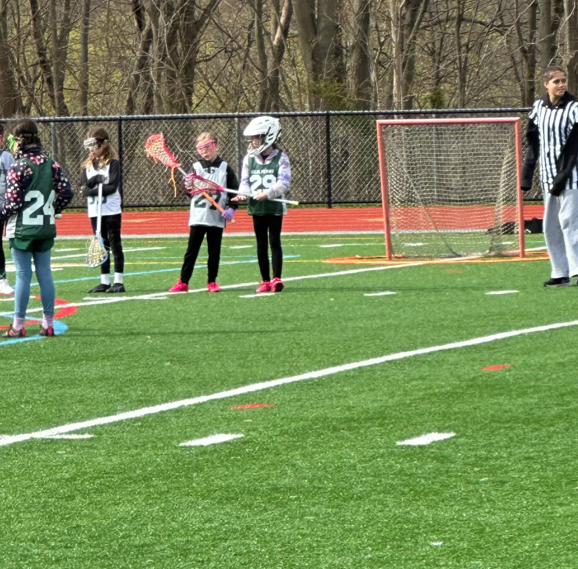 A wide shot capturing a youth girls' lacrosse game or practice on an outdoor green turf field with a red running track border