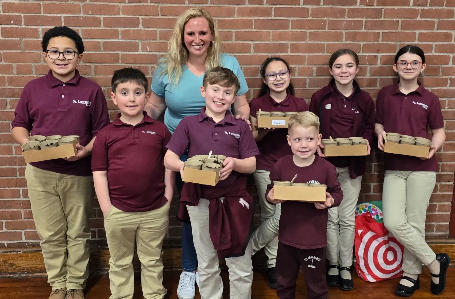 A group of seven young students in school uniforms and Shari-Lynn Cuomo Shore standing together smiling in front of a brick wall. The students are holding small cardboard trays containing small potted plants. Shari-Lynn stands behind the students in a blue t-shirt and jeans.