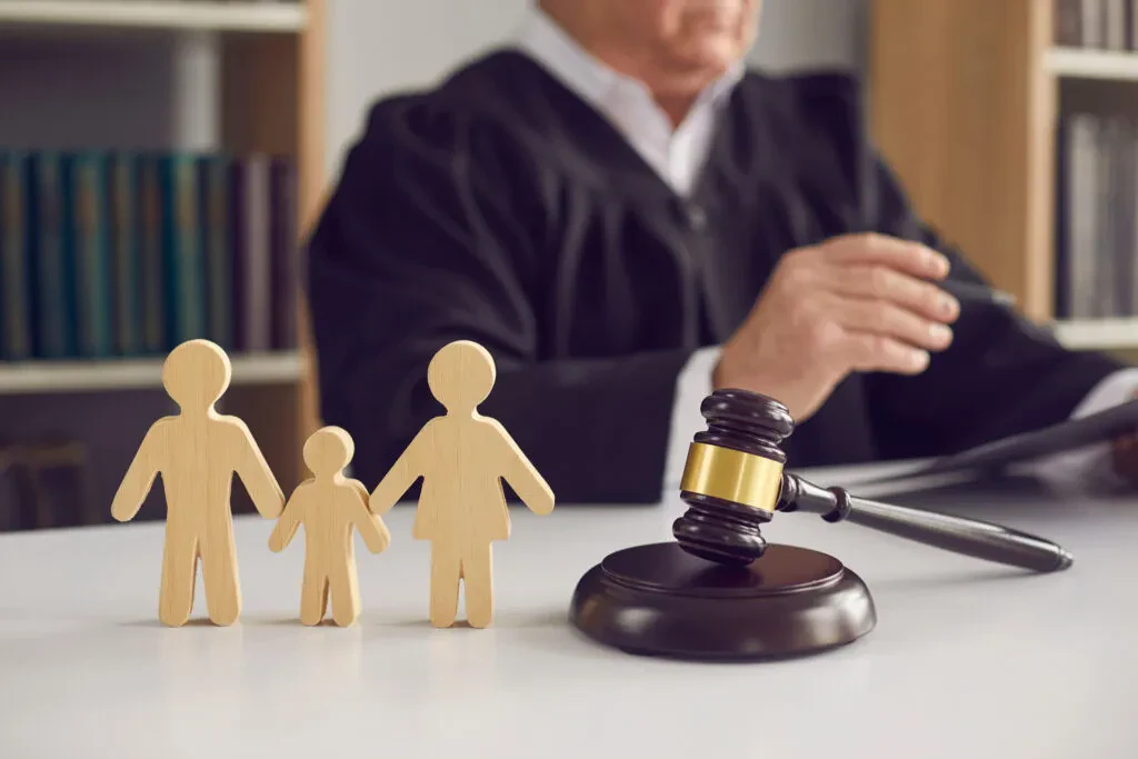 A judge sits at a desk with a gavel, behind wooden cutouts of two adults and one child, symbolizing family law or child custody decisions.