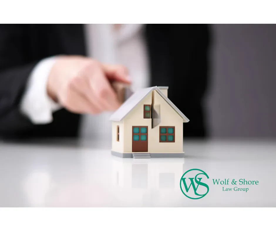 A small model house sits on a wooden cutting board, being sliced down the middle by a large kitchen knife held by an out-of-focus hand.