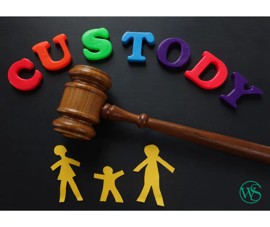 A judge’s gavel rests above paper cutouts of two adults and a child, with colorful letters spelling “CUSTODY” above them on a black background.