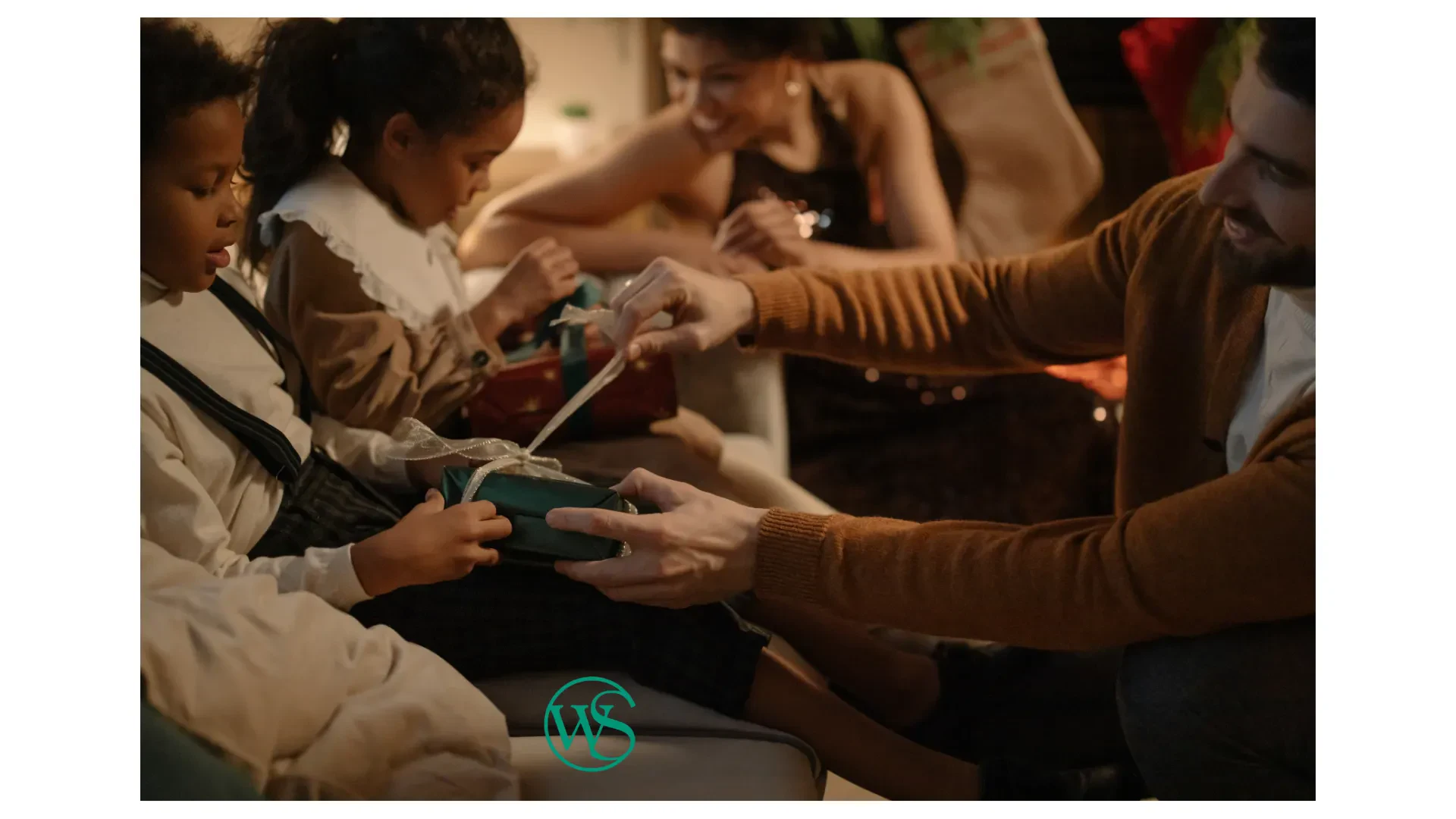 A man helps a child open a wrapped gift while another child and a woman watch, all sitting together in a cozy, festive room.