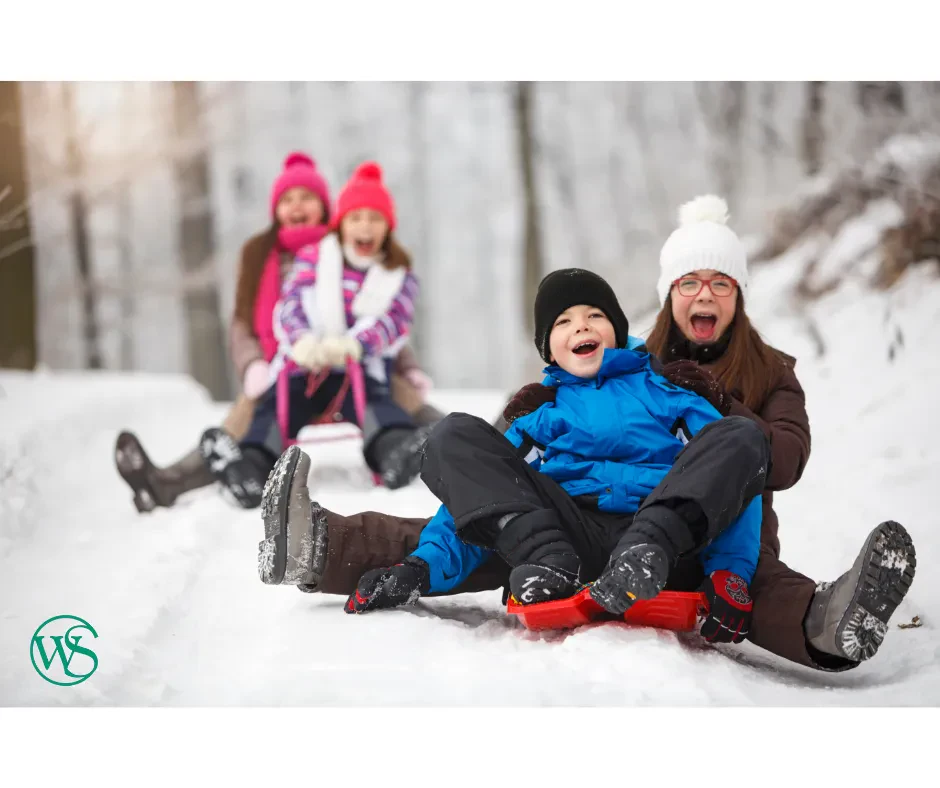Four children in winter clothes sled down a snowy hill, laughing and smiling. Snow-covered trees are in the background. The children are enjoying a fun day outside together.