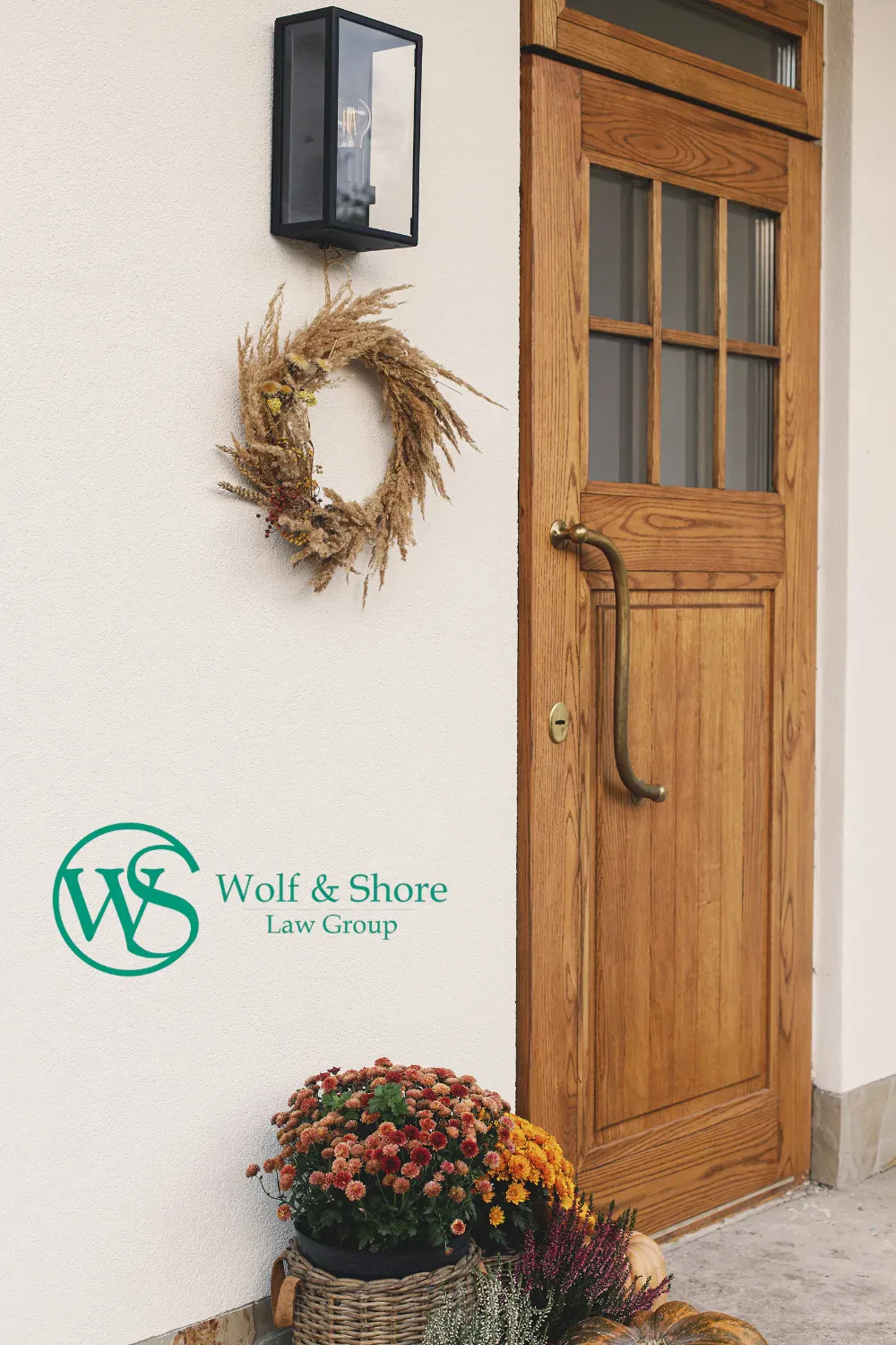 A wooden door with a wreath, outdoor light, and basket of flowers. “Wolf & Shore Law Group” with the WS logo is on the light-colored wall next to the door.
