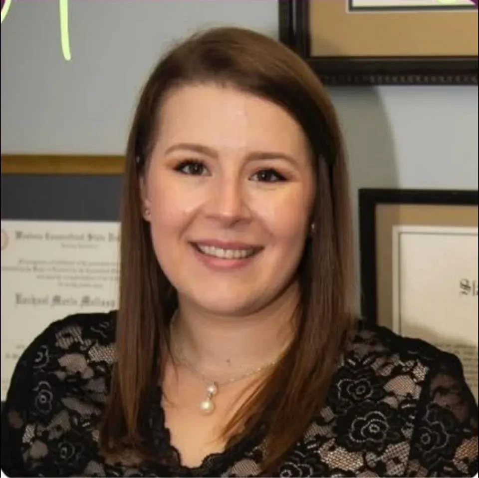 A medium close-up portrait of Rachael M. Levine with straight, shoulder-length brown hair parted to the side, smiling warmly at the camera. She is wearing a black lace-patterned top and a delicate silver necklace with two hanging pearl pendants.
