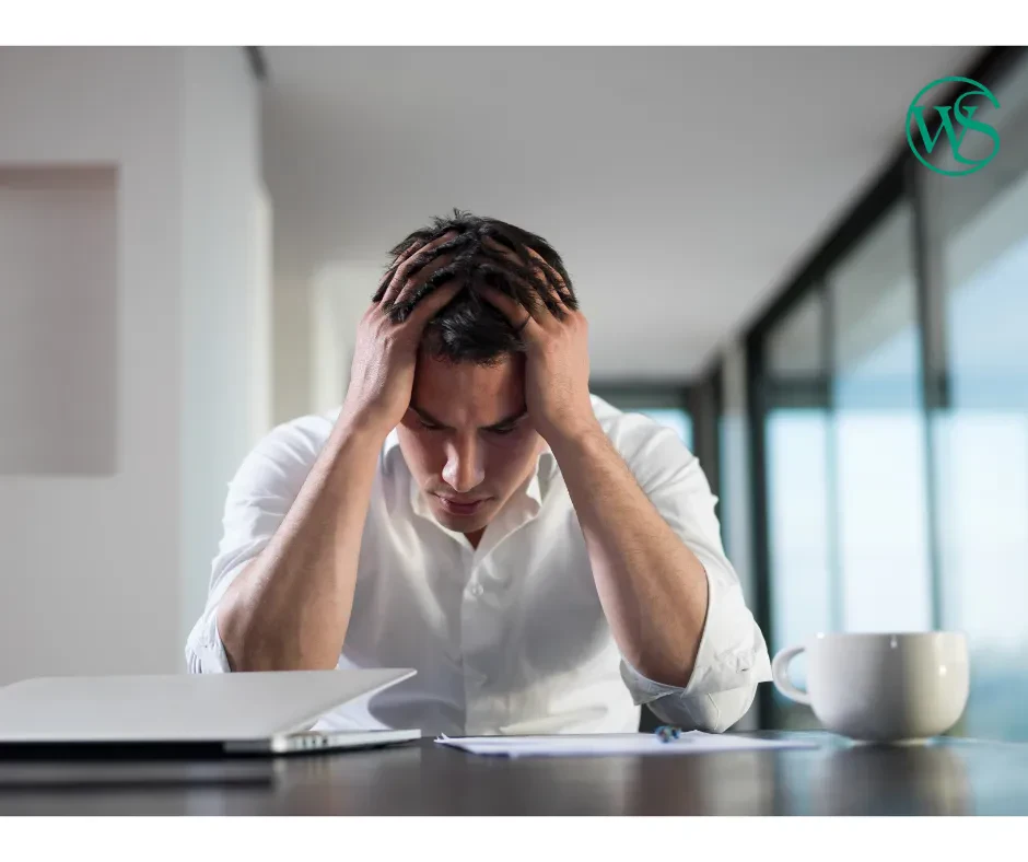 An image of a man in a white dress shirt sitting at a desk with his head in his hands, looking down in distress or exhaustion, with a laptop, paper, and a coffee mug in front of him.