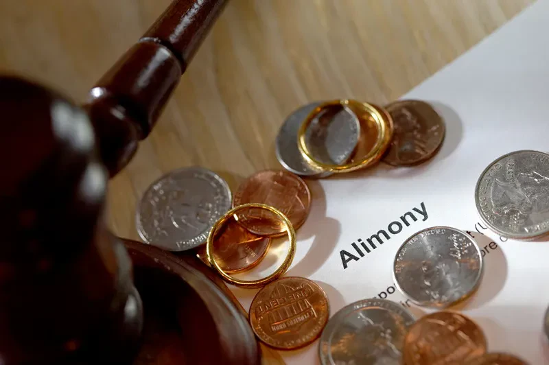 An image featuring a wooden judge's gavel on the left, with wedding rings, US coins, and a legal document clearly displaying the word "Alimony" scattered across the center and right.